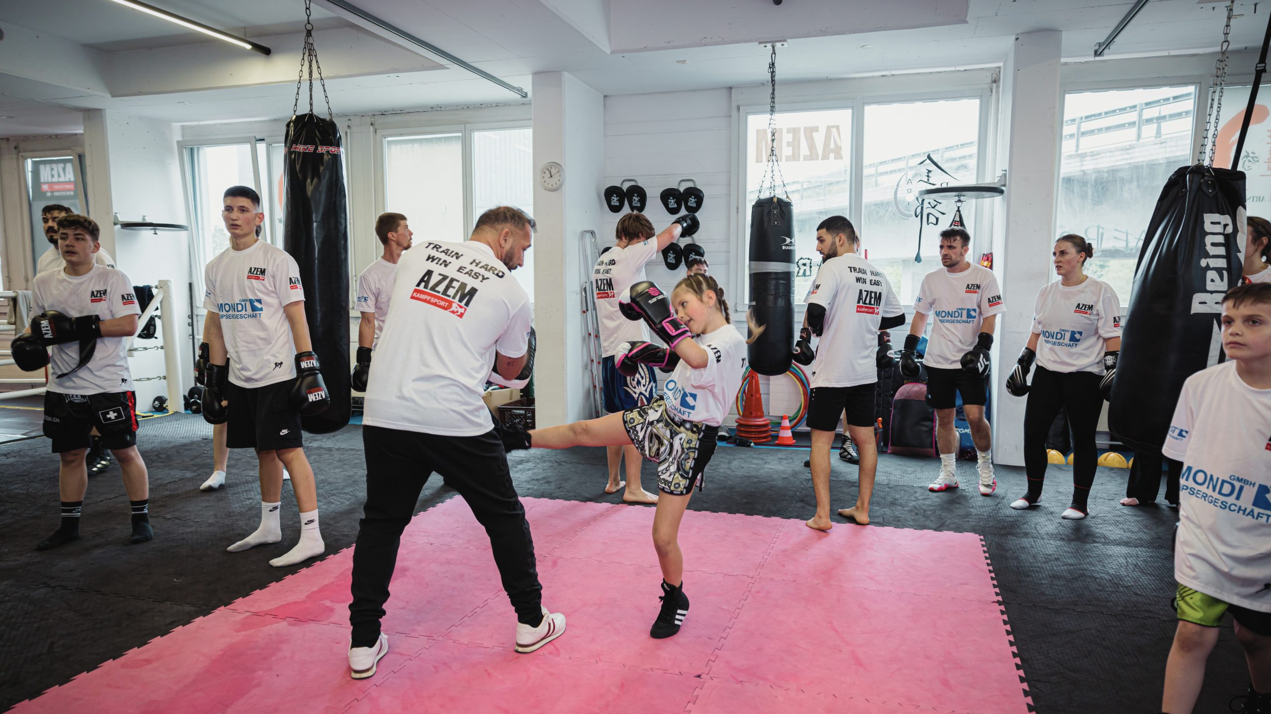 Junges Mädchen tritt im Kampfsporttraining bei AZEM gegen einen Trainer, umgeben von weiteren Teilnehmern