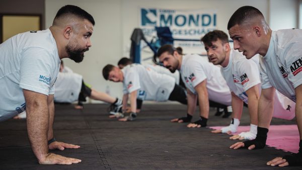 Kampfsporttrainer motiviert eine Gruppe junger Männer bei intensiven Liegestütz-Übungen im AZEM Gym
