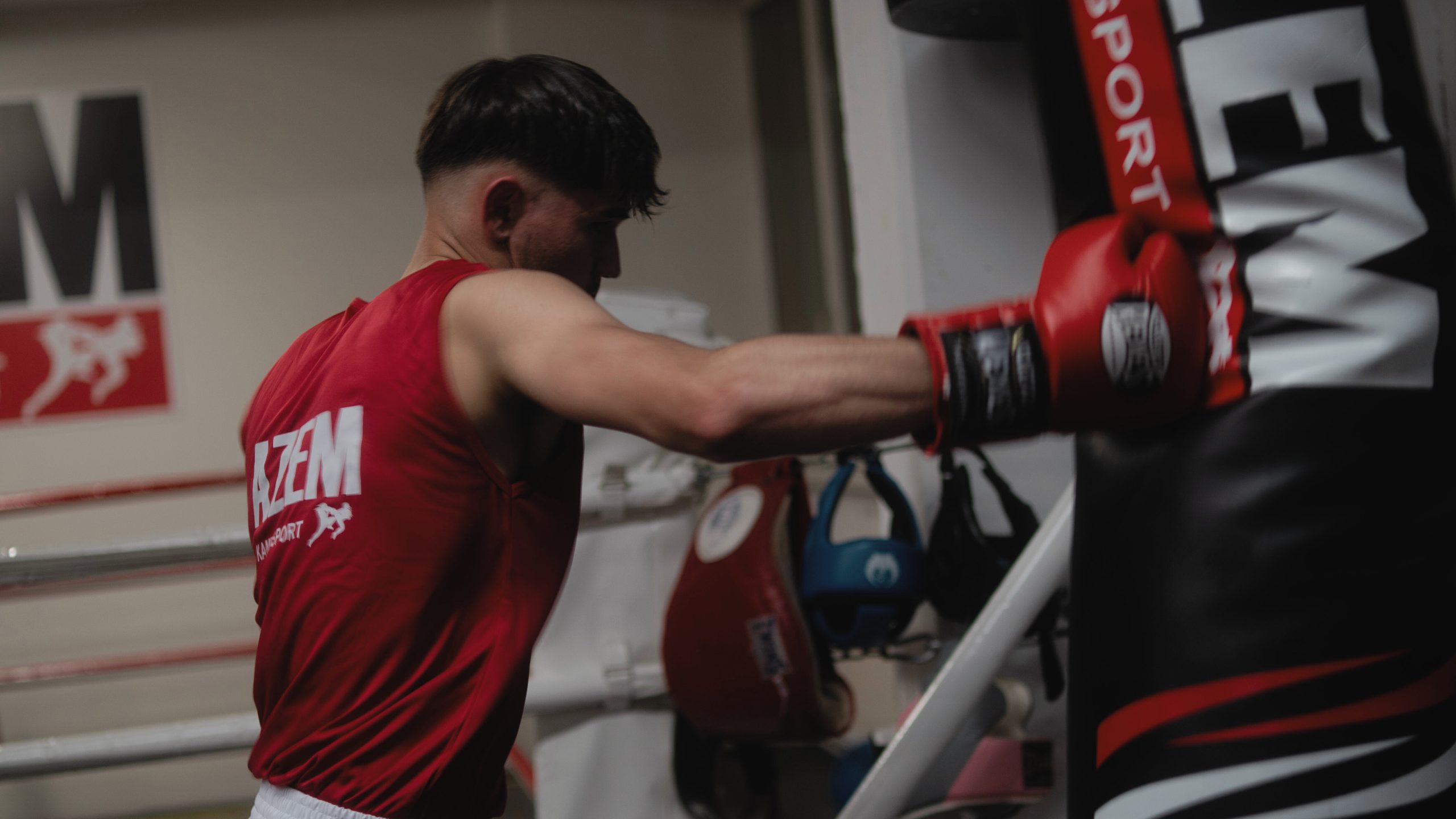 Boxer im roten AZEM Trikot schlägt mit roten Handschuhen kraftvoll gegen einen Sandsack im Trainingsraum